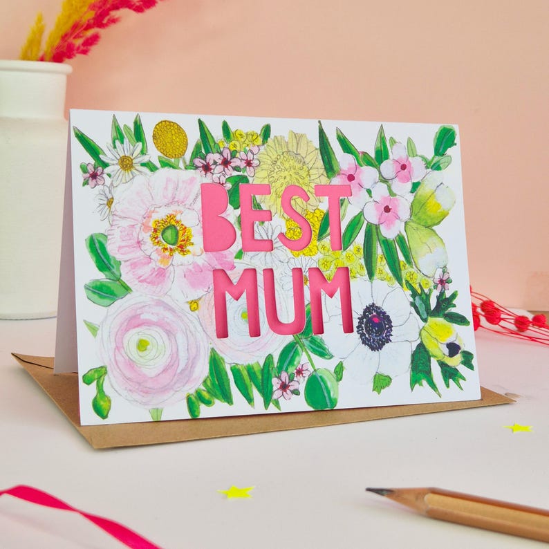 May include: A rectangular greeting card featuring a watercolor illustration of various flowers in shades of pink, yellow, and green. The text BEST MUM is die-cut in bold pink capital letters in the center of the floral arrangement on a white background.