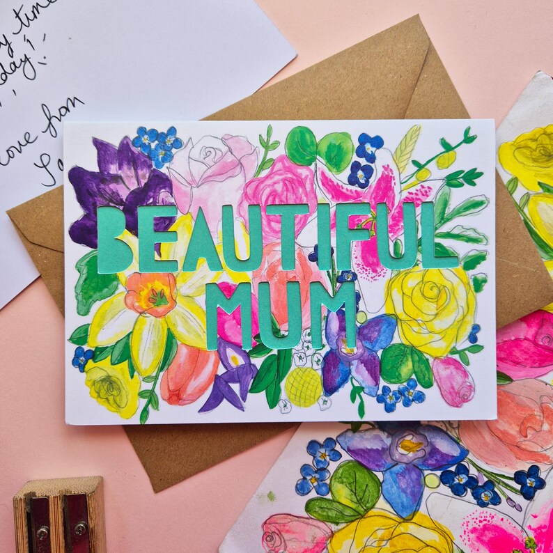 May include: A rectangular greeting card featuring a watercolor floral illustration in shades of pink, yellow, purple, and green. The text "BEAUTIFUL MUM" is die-cut in large, bold, teal capital letters across the center of the floral design.