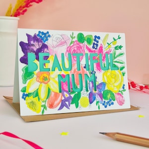 May include: A white greeting card featuring a vibrant watercolor floral illustration in shades of pink, yellow, purple, and green. The text BEAUTIFUL MUM is die-cut into the center of the card, revealing a solid teal background behind it.