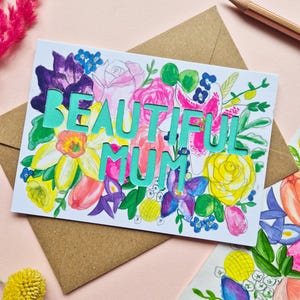 May include: A rectangular greeting card featuring a vibrant watercolor floral illustration in shades of pink, yellow, purple, and green. The words BEAUTIFUL MUM are die-cut in a bold, light teal font across the center of the floral design.