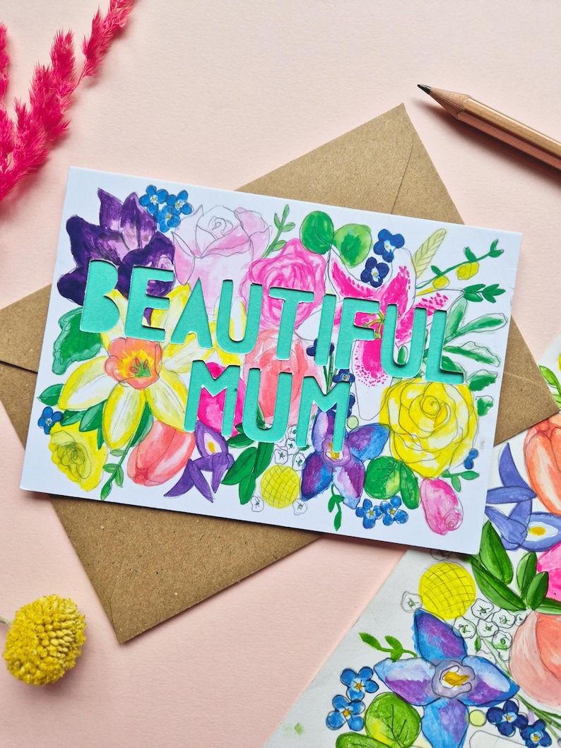 May include: A rectangular greeting card featuring a vibrant watercolor floral illustration in shades of pink, yellow, purple, and green. The words BEAUTIFUL MUM are die-cut in a bold, light teal font across the center of the floral design.