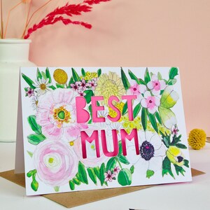 May include: A white greeting card featuring a watercolor illustration of various flowers in shades of pink, yellow, and green. The words BEST MUM are die-cut into the center of the card, revealing a bright pink layer underneath the floral design.