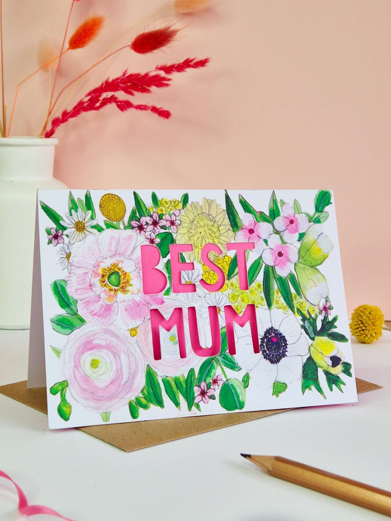 May include: A white greeting card featuring a watercolor illustration of various flowers in shades of pink, yellow, and green. The words BEST MUM are die-cut into the center of the card, revealing a bright pink layer underneath the floral design.