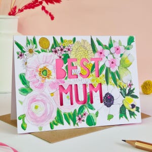 May include: A rectangular greeting card featuring a watercolor floral illustration with pink, yellow, and white flowers and green leaves. The words BEST MUM are die-cut into the center of the card, revealing a bright pink layer underneath.