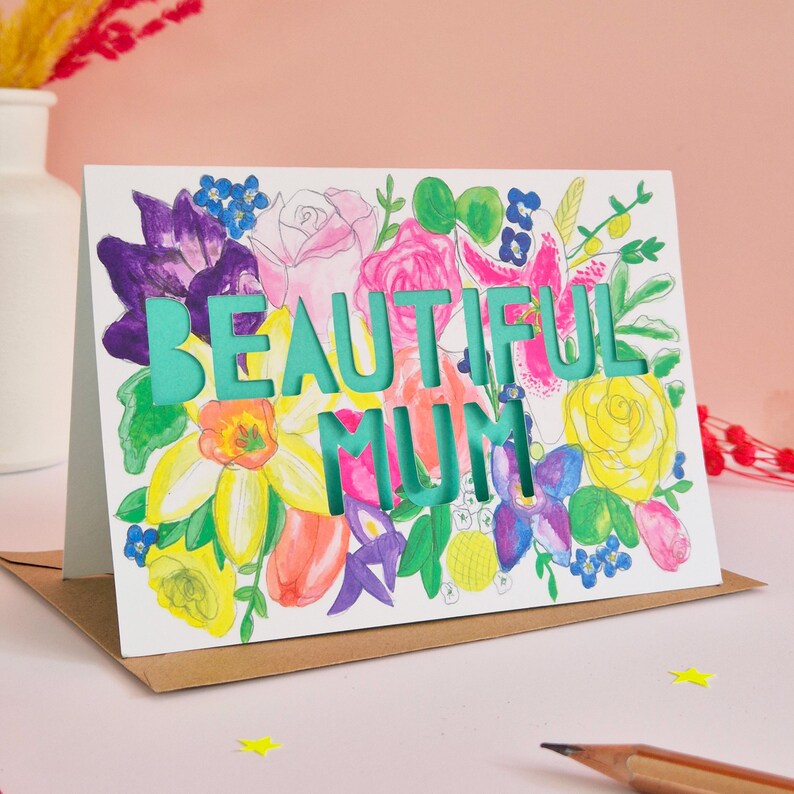 May include: A rectangular greeting card featuring a vibrant watercolor floral illustration with purple, pink, yellow, and green flowers. The words BEAUTIFUL MUM are die-cut into the center of the card, revealing a solid teal background layer.