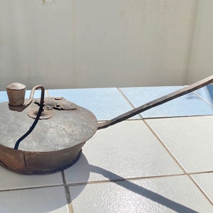 May include: A vintage, rusty metal oil lamp with a long handle and a small, round reservoir. The lamp has a wick and a spout for pouring oil.