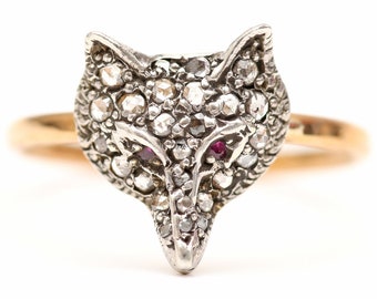 May include: A silver fox head ring with diamond accents and ruby eyes. The ring is set on a gold band.