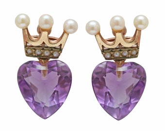 May include: A pair of earrings featuring heart-shaped amethyst stones. Each earring has a gold-colored crown design with small pearls. The earrings are set against a white background, highlighting the jewelry's details.