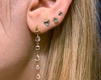 May include: A long, delicate earring with a row of small, triangular, clear gemstones. The earring is worn in the lower lobe of the ear. The ear also has three smaller, triangular, clear gemstones in the upper lobe and cartilage.
