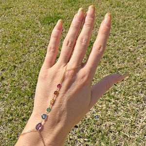 May include: A gold-colored hand chain featuring a series of colorful gemstones. The gemstones include red, orange, yellow, green, blue, and purple. The chain connects to a ring and drapes across the back of the hand.