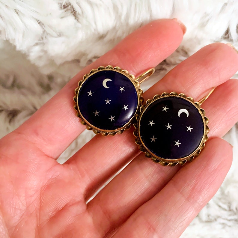 May include: A pair of gold-toned earrings with a dark blue enamel background and a white crescent moon and stars design.