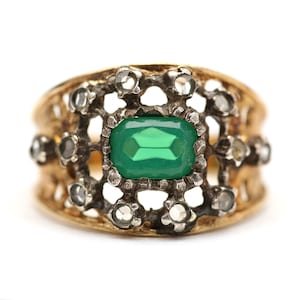 May include: A gold-toned ring with an emerald-cut, bright green gemstone at its center. The ring features a filigree design and is accented with small, clear gemstones around the central stone.