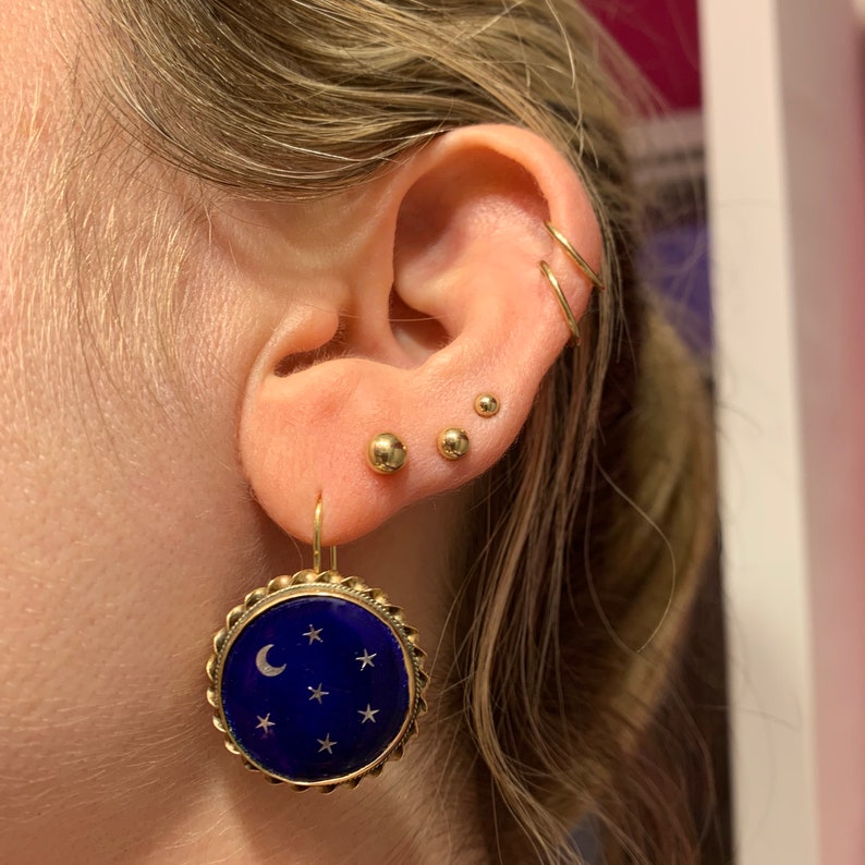 May include: Gold hoop earrings with a blue and gold star and crescent moon dangle earring. The dangle earring is a round disc with a blue enamel background and gold stars and a crescent moon.
