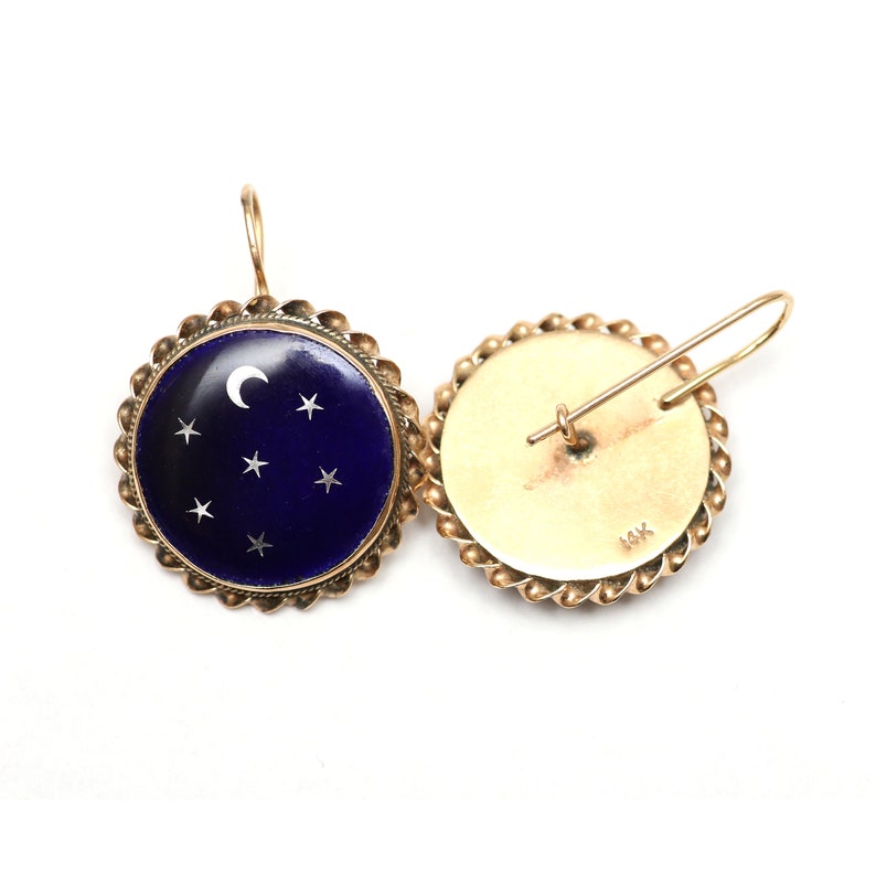 May include: A pair of gold earrings with a blue enamel and white enamel crescent moon and stars design. The earrings are marked "14K" on the back.