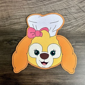 May include: A yellow and orange felt cutout of a dog wearing a white chef's hat with a pink bow. The dog has large brown eyes and a happy expression.