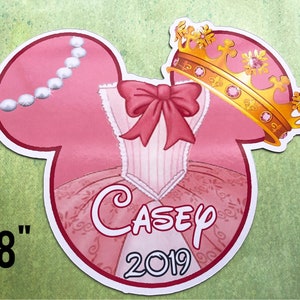 May include: Pink and white Mickey Mouse ears with a princess dress design, a gold crown, and the name "Casey" and the year "2019" printed on the ears. The ears are 8 inches tall.