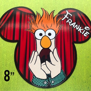 May include: A red and black Mickey Mouse-shaped cutout with a red curtain backdrop. The cutout features a cartoon character with orange hair, a white face, and a black beak. The character is holding its hands to its mouth in a surprised expression. The text "FRANKIE" is written in black on the cutout. The cutout is 8 inches tall.