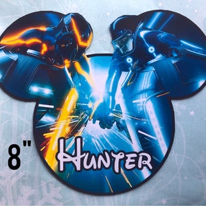 Peut inclure: Un autocollant en vinyle en forme de Mickey Mouse de 8 pouces représentant une scène du film Tron : L'Héritage. L'autocollant représente deux personnages conduisant des light cycles dans un schéma de couleurs bleu et orange. Le texte "Hunter" est imprimé sur l'autocollant.