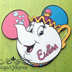 May include: A white cartoon teapot with a smiling face, a gold handle, and a purple and gold crown. The teapot has pink and blue Mickey Mouse ears. The name "Elaina" is written in pink script on the teapot. The text "sugah N spice" is at the bottom.