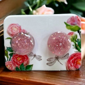 May include: A pair of pink glitter stud earrings on a white card with a floral design. The earrings are round and have a clear, shiny finish.