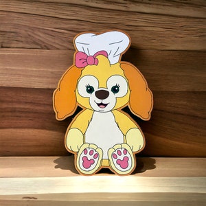 May include: A yellow plush toy dog wearing a white chef's hat with a pink bow. The dog has brown eyes, a black nose, and a pink tongue sticking out. The dog is wearing a white apron and has pink paw prints on its feet.