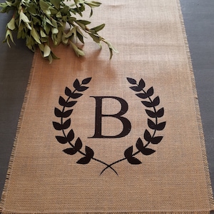 May include: A natural burlap table runner with a black laurel wreath design and the letter "B" in the center. The runner has a frayed edge and is decorated with a bundle of green leaves.