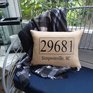 May include: A brown burlap pillow with black text that reads "29681 Simpsonville, SC". The pillow is sitting on a blue chair with a black and white plaid blanket.