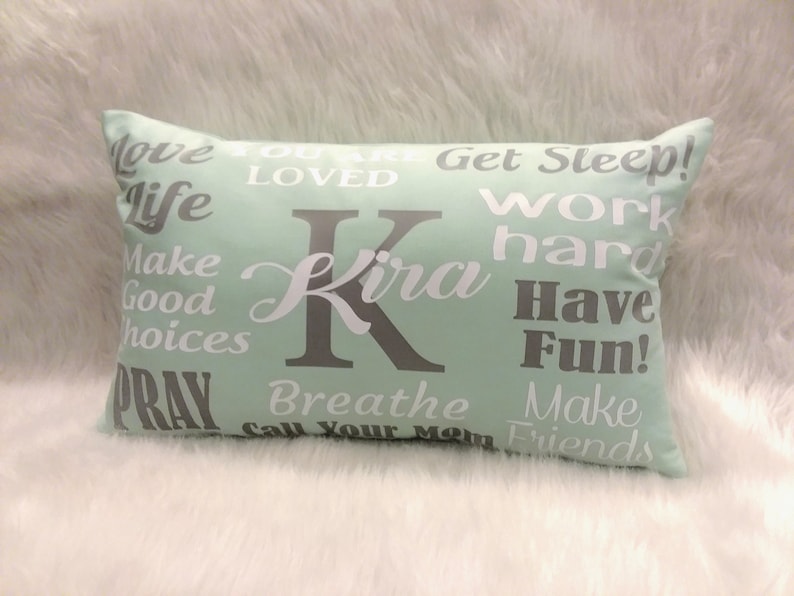 May include: A light green rectangular pillow with white and gray text. The text says "Love Life", "You Are Loved", "Get Sleep", "Work Hard", "Make Good Choices", "Pray", "Breathe", "Call Your Mom", "Make Friends", "Have Fun!" and "Kira".
