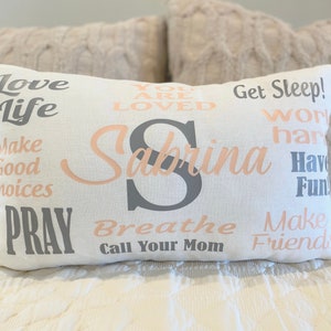 May include: A white pillow with the name Sabrina in pink lettering and various inspirational phrases in gray and pink lettering, including "Love Life", "Get Sleep", "Work Hard", "Have Fun", "Make Good Choices", "Pray", "Breathe", "Call Your Mom", and "Make Friends".