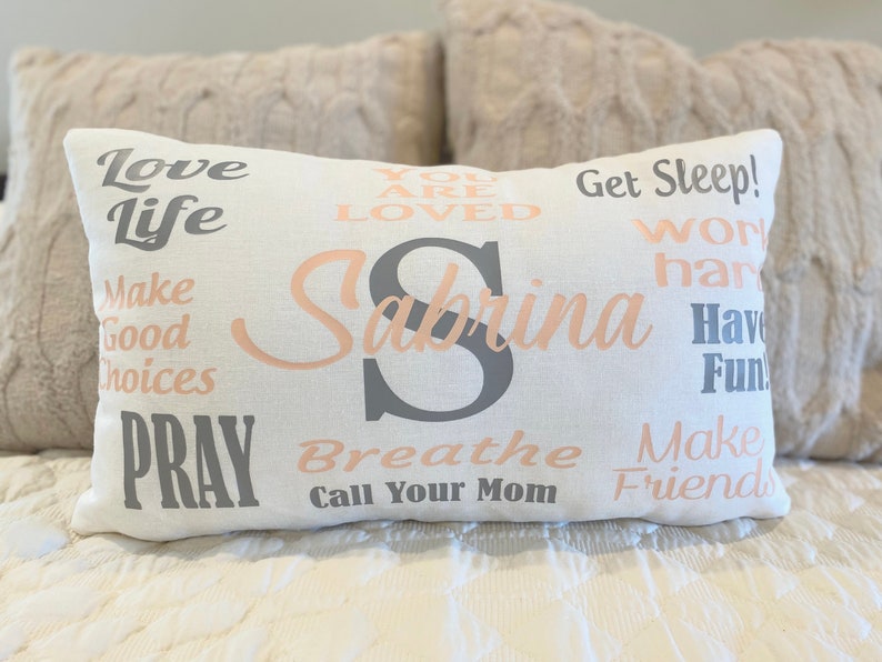 May include: A white pillow with the name Sabrina in pink lettering and various inspirational phrases in gray and pink lettering, including "Love Life", "Get Sleep", "Work Hard", "Have Fun", "Make Good Choices", "Pray", "Breathe", "Call Your Mom", and "Make Friends".