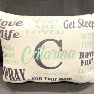 May include: White pillow with a green and black design. The pillow has the text "Love Life", "You Are Loved", "Get Sleep", "Work Hard", "Make Good Choices", "Have Fun", "Pray", "Breathe", "Call Your Mom", "Make Friends", and "Catarina" on it.