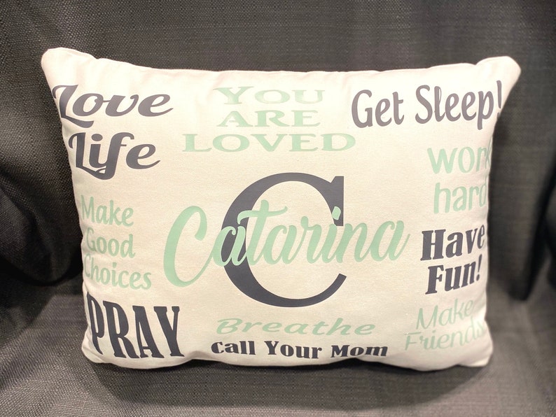 May include: White pillow with a green and black design. The pillow has the text "Love Life", "You Are Loved", "Get Sleep", "Work Hard", "Make Good Choices", "Have Fun", "Pray", "Breathe", "Call Your Mom", "Make Friends", and "Catarina" on it.