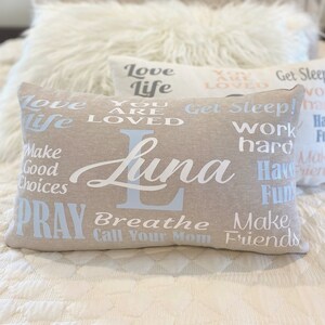 May include: A beige pillow with the name "Luna" printed in white cursive lettering. The pillow is decorated with various phrases in white and light blue lettering, including "Love Life", "You Are Loved", "Get Sleep", "Work Hard", "Have Fun", "Make Good Choices", "Pray", "Breathe", "Call Your Mom", and "Make Friends".