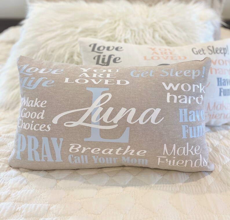 May include: A beige pillow with the name "Luna" printed in white cursive lettering. The pillow is decorated with various phrases in white and light blue lettering, including "Love Life", "You Are Loved", "Get Sleep", "Work Hard", "Have Fun", "Make Good Choices", "Pray", "Breathe", "Call Your Mom", and "Make Friends".