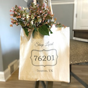 May include: A white canvas tote bag with the text "Shop Local" and the number "76201" in a decorative frame. The text "Denton, TX" is printed below the frame. A bouquet of pink flowers is in the bag.