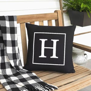 May include: A black square pillow with a white border and a large white letter "H" in the center. The pillow is on a wooden bench with a black and white checkered throw blanket. A decorative item for home decor.