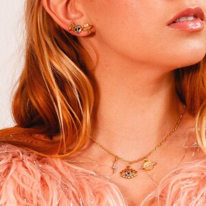 May include: Gold necklace with a charm of an eye with colorful gemstones, a lightning bolt, and a planet. The necklace is worn by a person with long red hair.