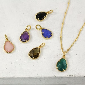 May include: A set of six gold-tone teardrop-shaped pendants with different colored gemstones. The pendants are on a white surface.
