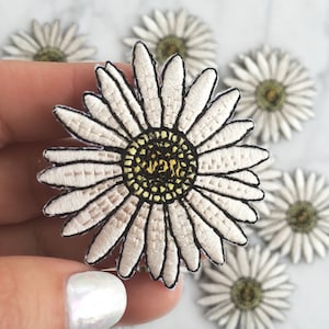 May include: White embroidered daisy flower patch with a yellow center. The patch is surrounded by other daisy patches.