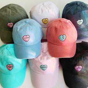 May include: Nine different colored baseball caps with candy heart embroidery. The caps are in various colors including black, white, pink, blue, green, and red. The candy hearts have different sayings on them, including "WTF", "Bye", "Nah", "Love", "As If", "Thank U, Next", "Bored", "Bad Bitch".