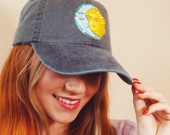 May include: A dark blue denim baseball cap with a sun and moon embroidery design. The embroidery is in yellow, white, and blue. The cap is worn by a person with long red hair.