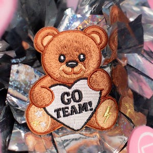 May include: A brown teddy bear patch holding a white heart with the text "GO TEAM!" and a lightning bolt. The patch is surrounded by silver and pink pom-poms and pink foam fingers.