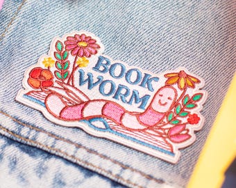 Könnte beinhalten: Hellblaue Jeansjacke mit einem farbenfrohen, gestickten Aufnäher. Der Aufnäher zeigt einen lächelnden, rosa Wurm mit Blumen und dem Text "BOOK WORM". Bücher in Rosa und Gelb sind im Hintergrund zu sehen.