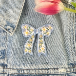 以下が含まれることがあります： デニムジャケットの上に、デイジーの花柄が施された水色のリボン型のピン。ピンには白と黄色のデイジーの繊細な模様があります。ピンクと白のチューリップが2本、背景にあります。