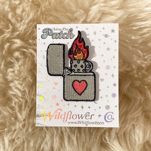 May include: Iron-on patch featuring a gray lighter with a red heart and a flame. The patch is on a white card with the text "Iron-on Patch" and "Wildflower + Co. www.Wildflowerco.com".