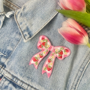 以下が含まれることがあります： イチゴのデザインが施されたピンク色のリボン型のパッチ。パッチは、ピンク色の背景に赤いイチゴと白い花が特徴です。パッチは、薄い青色のデニムジャケットの上に配置され、背景にはピンク色のチューリップがあります。