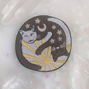 May include: Embroidered patch featuring two cats, one white and one black, curled up together in a circle. The white cat is facing the sun, while the black cat is facing the moon. The patch is surrounded by stars and has a brown background.