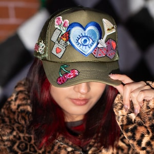 Puede incluir: Gorra de camionero verde oliva con parches bordados. Los parches incluyen un corazón azul con un ojo, cerezas, dados, un encendedor y flores. La gorra tiene una parte trasera de malla y una visera curva.