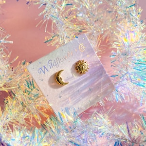 May include: A pair of gold earrings featuring a crescent moon and a sun with a face. The earrings are on a white card with the text "Wildflower Co."