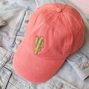 Peut inclure: Une casquette de baseball rouge avec un cactus brodé. Le cactus est vert avec des fleurs jaunes et est centré sur le devant de la casquette.
